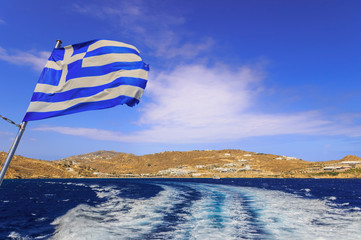 Discovering the Greek islands. Horizon with ship wake and the Greek flag: island of Mykonos in the...