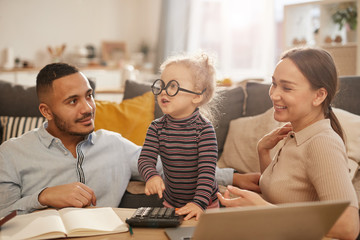 Portrait of happy young family counting home finances with cute little girl wearing glasses in...