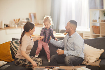 Warm toned portrait of happy modern family playing with cute little daughter in sunlit home interior, copy space