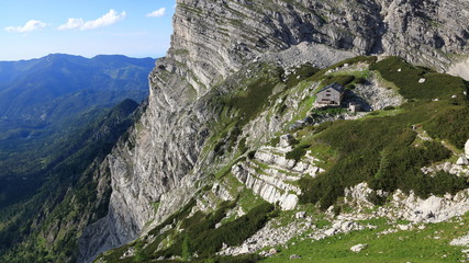 New Alpine hut on green grass surrounded by mighty rocks with forested mountain ridge in the background, Alps, Austria.