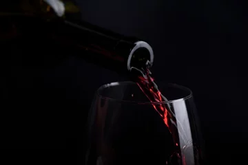Gordijnen Red wine pouring in wine glass over black background. Closeup of red wine splashing in wineglass in restaurant. © hitdelight