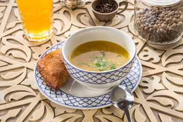 chicken broth with bread bun and parsley in bowl with glass of yellow drink on oriental wooden table