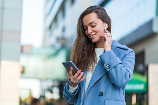 Stylish trendy happy joyful brunette woman in a blue coat holds a smartphone and using wireless white headphones for listen music in the city centre. Modern people