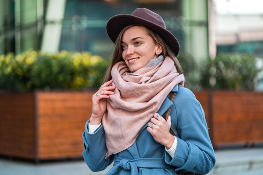 Beautiful happy smiling stylish trendy brunette hipster woman in a brown hat and in a blue coat with cozy warm scarf walking around the city. Fashionable people