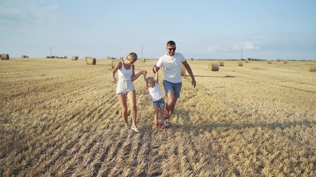 Happy parents hold daughter hands, run and throw hay in huge summer field