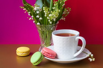 French multicolored cakes macarons and a cup of tea. A bouquet of lily of the valley in a crystal vase.