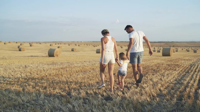 Back view of happy parents walk with their daughter for a hand in field