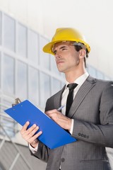Young male architect writing on clipboard while looking away outside office building