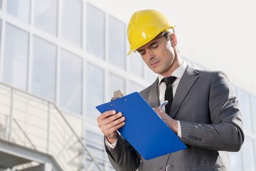 Young male architect writing on clipboard against office building