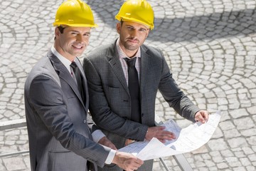 Portrait of confident young businessmen in hard hats examining blueprint outdoors