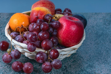 red grape berries and fresh fruits in a straw basket on a dark wooden background.