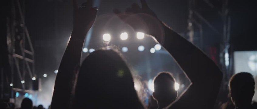 Behind shot of people crowd waving hands, jumping and dancing in front of a stage during a live show concert or a night club, light show at the background. Medium, back shot, slow motion. BMPCC 4K
