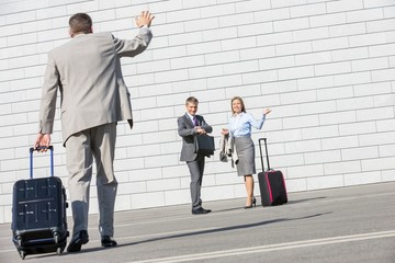 Rear view of businessman carrying luggage waving hand to colleagues