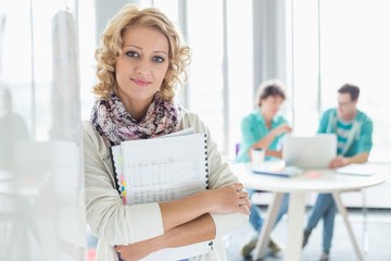 Portrait of creative businesswoman holding files with colleagues working in background at office