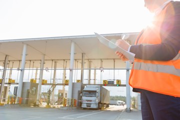 Midsection of female worker writing on clipboard while looking at truck entering in shipping yard