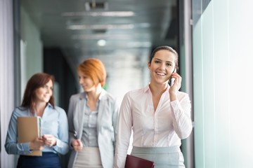 Young businesswoman on call with colleagues in background at office corridor