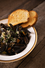 Mussels in white wine sauce with herbs in a bowl with golden croutons on a dark, wooden board.