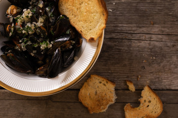Mussels in white wine sauce with herbs in a bowl with golden croutons on a dark, wooden board.