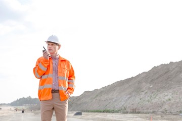 Confident male supervisor using walkie-talkie on construction site against clear sky