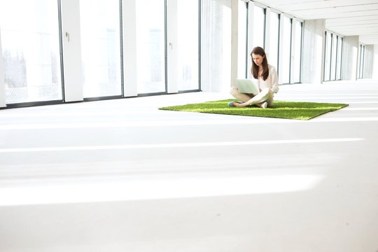 Young businesswoman using laptop while sitting on turf in empty office