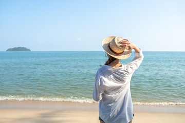 Fototapeta na wymiar Back view or behind view of beautiful young woman wearing white shirt and straw hat standing on beach with sea and sky background