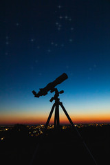 Telescope silhouette and night sky with city lights in the background.
