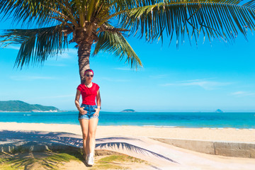 Beautiful pretty girl tourist, young woman is resting, standing under a palm tree in sunglasses and red T-shirt. Enjoying summer vacation, sea, beach tropical  background. Tourism concept.