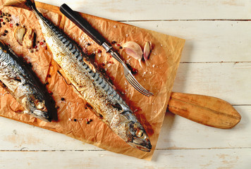 Baked mackerel with garlic, pepper, fork and coarse salt on baking paper and cutting board on white rustic wooden boards.