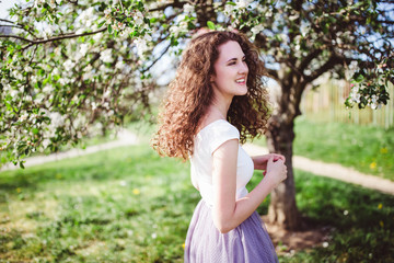 Fototapeta premium Happy curly hair young woman in a white t-shirt under the blooming apple tree, smiling.