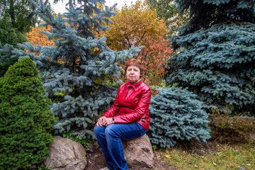 A lady in a red jacket and jeans sits on a stone against a background of trees