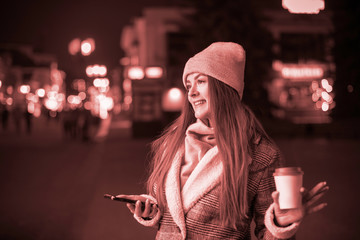 A young beautiful girl with long hair in a fashionable hat and coat, with a Cup of hot coffee and a phone on the background of the lights of the night city of bokeh in winter. Toning.