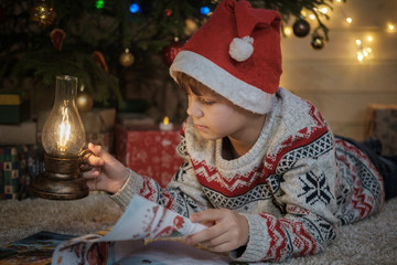 a cute boy in a red hat is reading a children's book near a Christmas tree and holding a vintage...