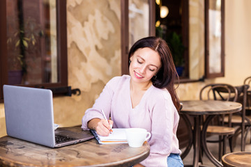 Fototapeta premium Smiling woman in cafe using laptop and note some data on notebook