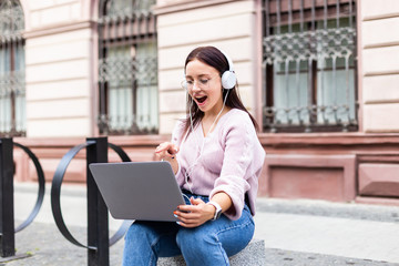 Young girl listening music with headphones and searching songs in a laptop sitting on a bench in the street