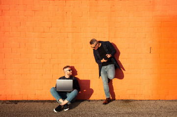 Two friends in casual-wear in urban contest with digital devices. Young caucasian man sitting cross-legged on ground and working on laptop. The other man standing with smartphone. Red wall background.