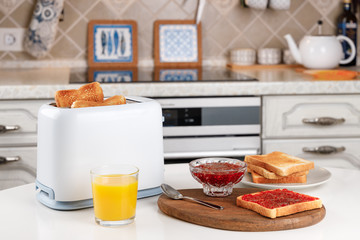 White toaster, glass of orange juice, slices of toasted bread, crispy toast with raspberry jam and bowl with confiture on wooden board. Served table for delicious meal. Fresh breakfast concept.
