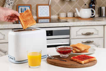Male hand takes the toaster bread out of the white toaster. Glass of orange juice, bowl with strawberry confiture, toast with raspberry jam. Delicious breakfast concept. Morning meal. Kitchen interior