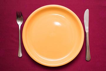 Empty yellow plate, fork and knife on maroon tablecloth. Elegant table setting. Top view, closeup