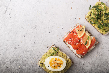 Three open sandwiches with avocado, red fish, egg and micro greenery. Light gray background.