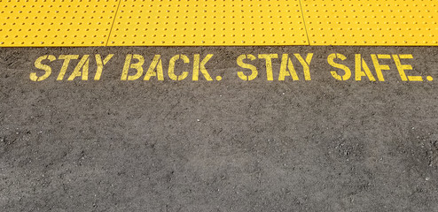 yellow sign at train station asking people to stay back, stay safe, on concrete floor
