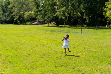 back view of brunnette young girl with white dress and red shoes running in green field near a forest, sun shining in spring