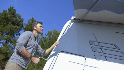 A man sets up a canopy from the sun on a motor home against a clear blue sky