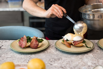 Eggs benedict with crisp bacon and spinach being topped with hollandaise sauce for morning brunch.