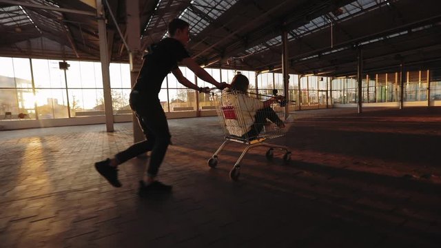 Crazy couple riding a supermarket trolley in an empty store, sun glare