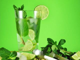 Glass of mojito or natural lemonade with lime slices, mint leaves and ice cubes next to peppermint sprigs, ice tongs and citrus pieces on light green background. 
