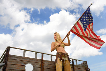A beautiful young American girl stands on an old cattle trailer. She proudly waves the US flag   cheerfully as the wind blows through her hair.