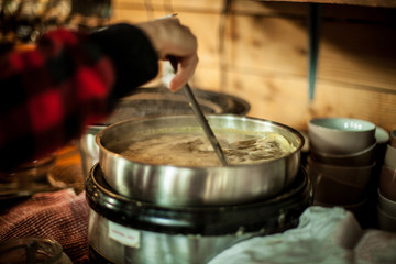 Side view close up of homemade healthy soup in big metallic pot. Hand is stirring the meal with scoop while it is boiling. Blurred kitchen and dishes in the background.