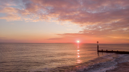 An aerial view of a beautiful sunset with orange circle sun and reflection on a calm sea along a sandy beach and groyne (breakwater) under a blue sky and orange clouds