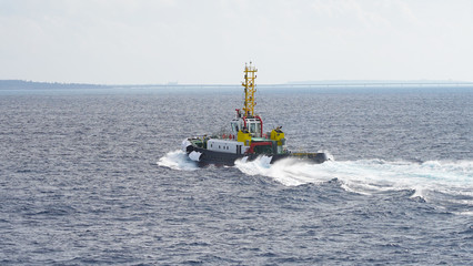 Fototapeta premium Colored tugboat works in port. bright tugboat in blue water. industrial ship on waves tropical islands. powerful tugboat helps maneuvering big ship turquoise blue Pacific Ocean Okinawa Japan