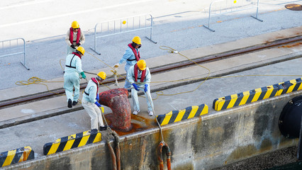 a group of Japanese port workers in uniform and orange helmets fasten ship mooring ropes to the bollard in the port. workers moor ship